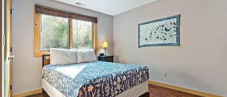 A bedroom with a bed, a window, a wall-mounted quilt, and a nightstand with a lamp.