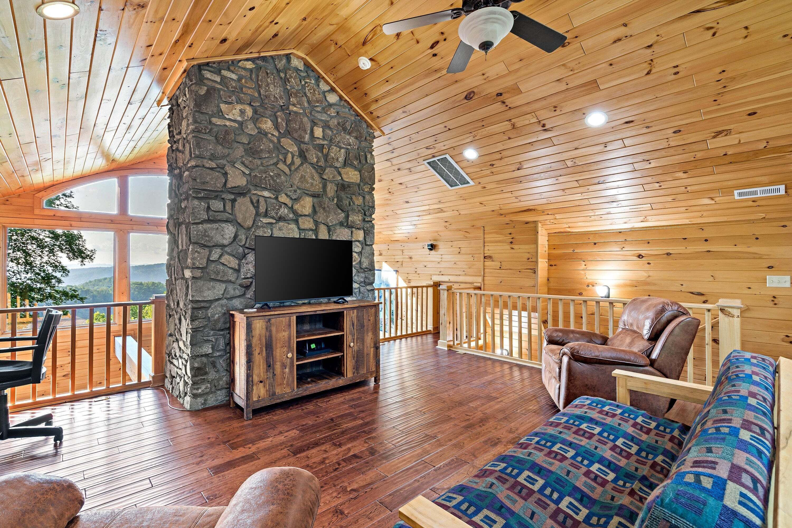 A cozy cabin interior with a stone fireplace, wooden flooring, a flat-screen TV, and a comfortable seating area.