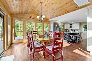 A wooden dining area with a large table and chairs, a kitchen with white cabinets, and a view of greenery outside.