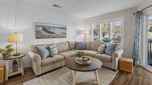 Una sala de estar moderna con un sofá seccional color beige, una mesa de centro redonda y un cuadro de un barco en la playa.
