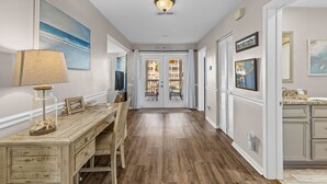 Un pasillo con un escritorio de madera, una lámpara y un cuadro enmarco. Una puerta con la etiqueta "Beach House" da a un balcón.