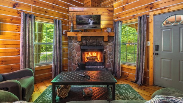 A log cabin interior with a stone fireplace, a television mounted above it, and a cozy seating area with a table. - Rustic Cabin Getaway with Hot Tub, Trails & Fire Pit (Logan)