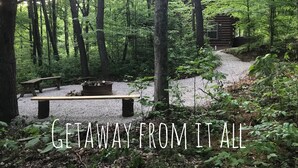 A forested area with a wooden cabin, a gravel path, and a bench. - Rustic Cabin Getaway with Hot Tub, Trails & Fire Pit (Logan)