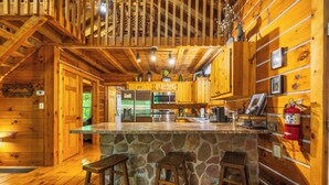 A wooden cabin interior with a stone island bar, wooden stools, a kitchen with appliances, and a fire extinguisher. - Rustic Cabin Getaway with Hot Tub, Trails & Fire Pit (Logan)