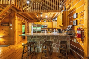 A wooden cabin interior with a stone island bar, wooden stools, a kitchen with appliances, and a fire extinguisher.