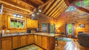 A wooden cabin with a kitchen, dining area, and a large window. - Rustic Cabin Getaway with Hot Tub, Trails & Fire Pit (Logan)