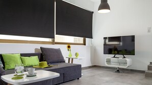 Ein modernes Wohnzimmer mit einem grauen Sofa, einem kleinen, weißen Tisch mit Glasplatte, einem Flachbildfernseher und schwarzen Jalousien an den Fenstern.