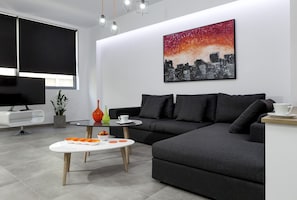Una sala moderna con un sofá seccional, un televisor de pantalla plana, una mesa de centro con fruta y un cuadro de paisaje urbano en la pared.