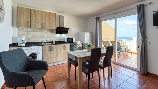 Cocina moderna con armarios de madera, microondas, cafetera y lavadora. Cuenta con comedor con mesa y sillas, además de un balcón con vistas al mar.