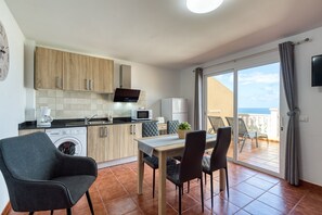Una cuina moderna amb armaris de fusta, microones, cafetera i rentadora. Hi ha una zona de menjador amb taula i cadires, i un balcó amb vista al mar.