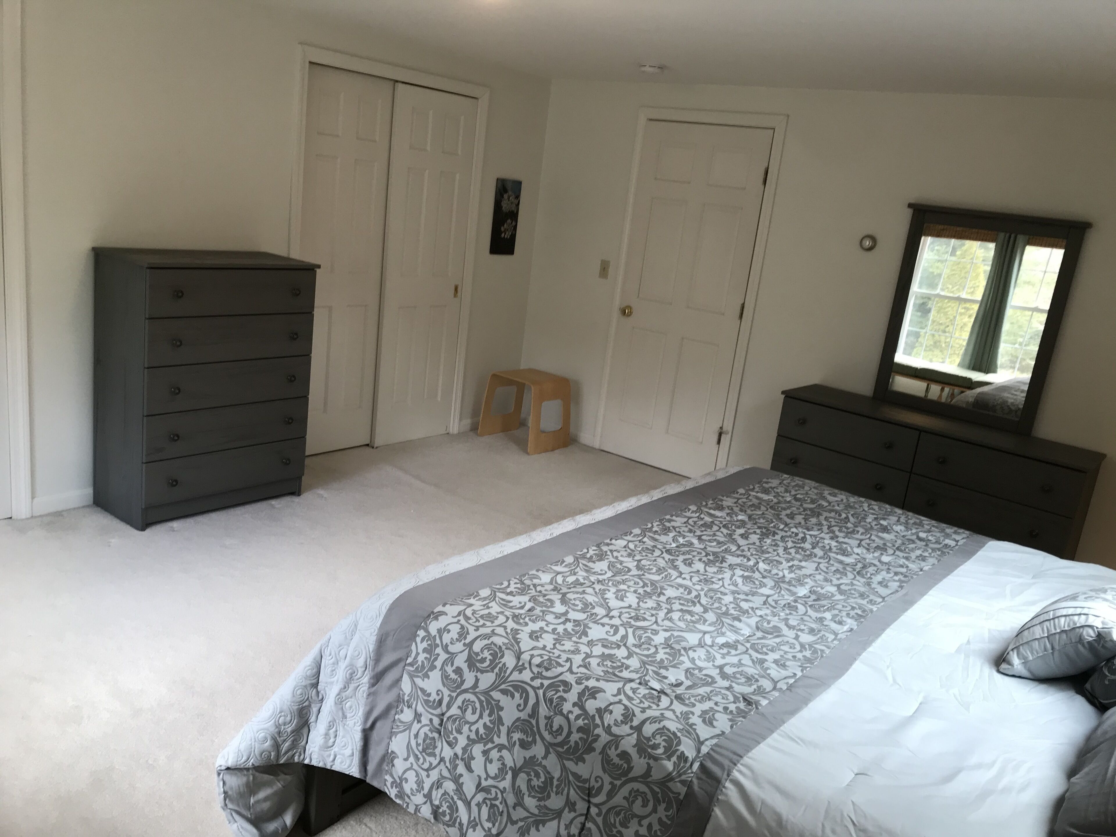 A bedroom with a bed, two dressers, a small stool, and a window with a view.