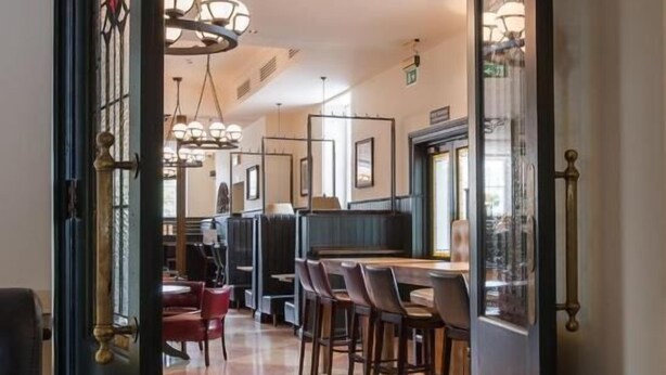 Un couloir mène à une salle à manger dont le sol est décoré de motifs, où l’on trouve des chaises en bois et un coin bar avec des tabourets.