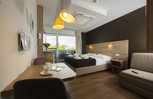 Quarto de hotel moderno com piso de madeira, uma cama com cabeceira, um sofá, uma mesa de jantar com xícaras, um abajur e vista para o exterior.