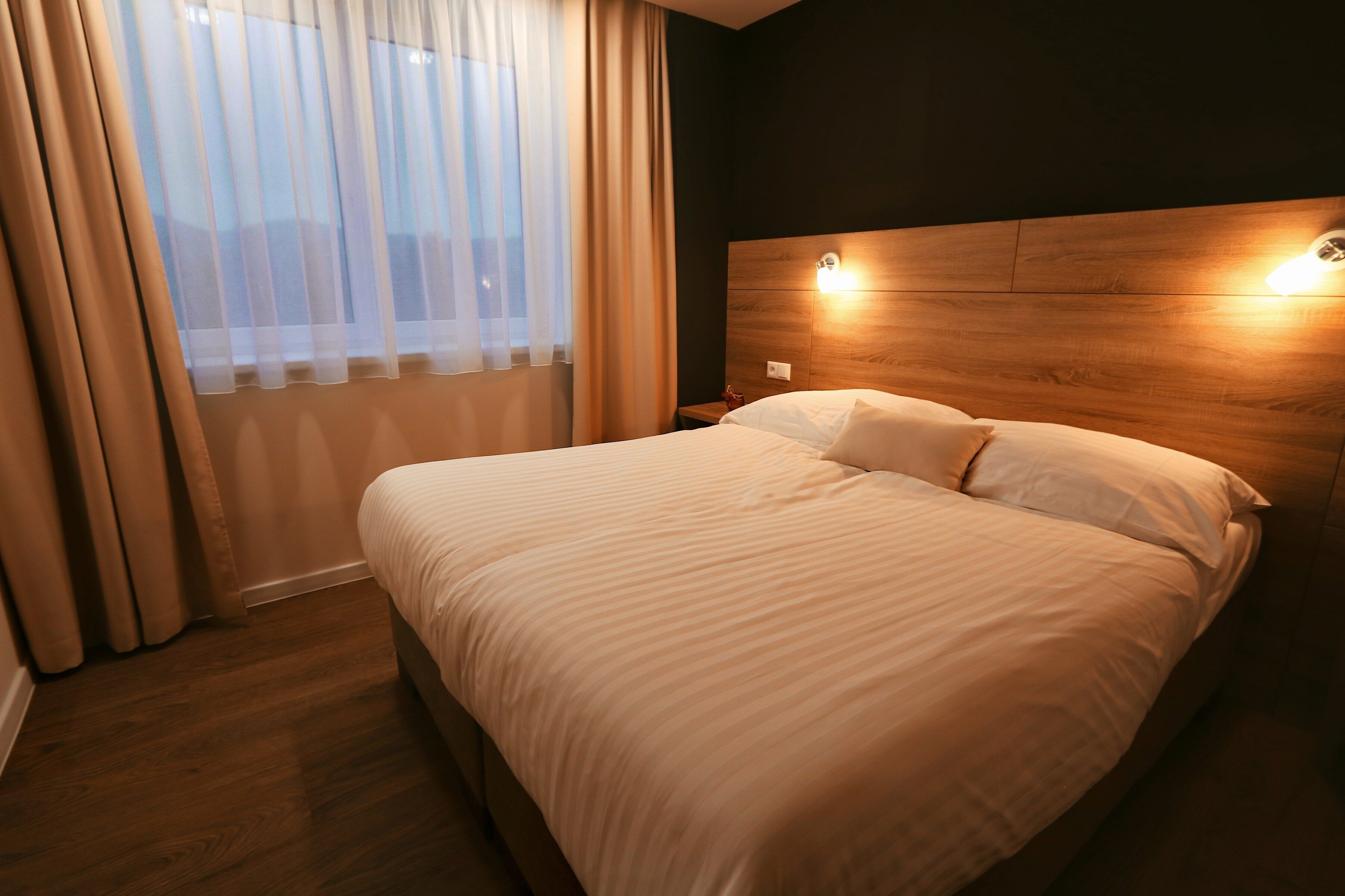 A hotel room with a large bed, wooden headboard, bedside lamps, and a window with sheer curtains.