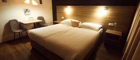 A hotel room with a large bed, a desk with two chairs, and a wooden headboard with integrated lighting.