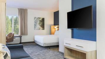 Habitación de hotel con cama, sofá, escritorio con silla, televisor de pantalla plana montado en la pared y ventana con cortinas.