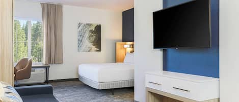 A hotel room with a bed, a sofa, a desk with a chair, a flat-screen TV mounted on the wall, and a window with curtains.
