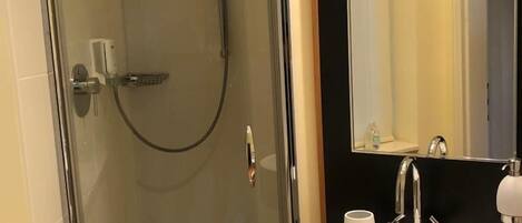 Una ducha envuelta en vidrio con cabezal de ducha manual, un lavamanos blanco con un solo grifo y un espejo grande con marco negro.
