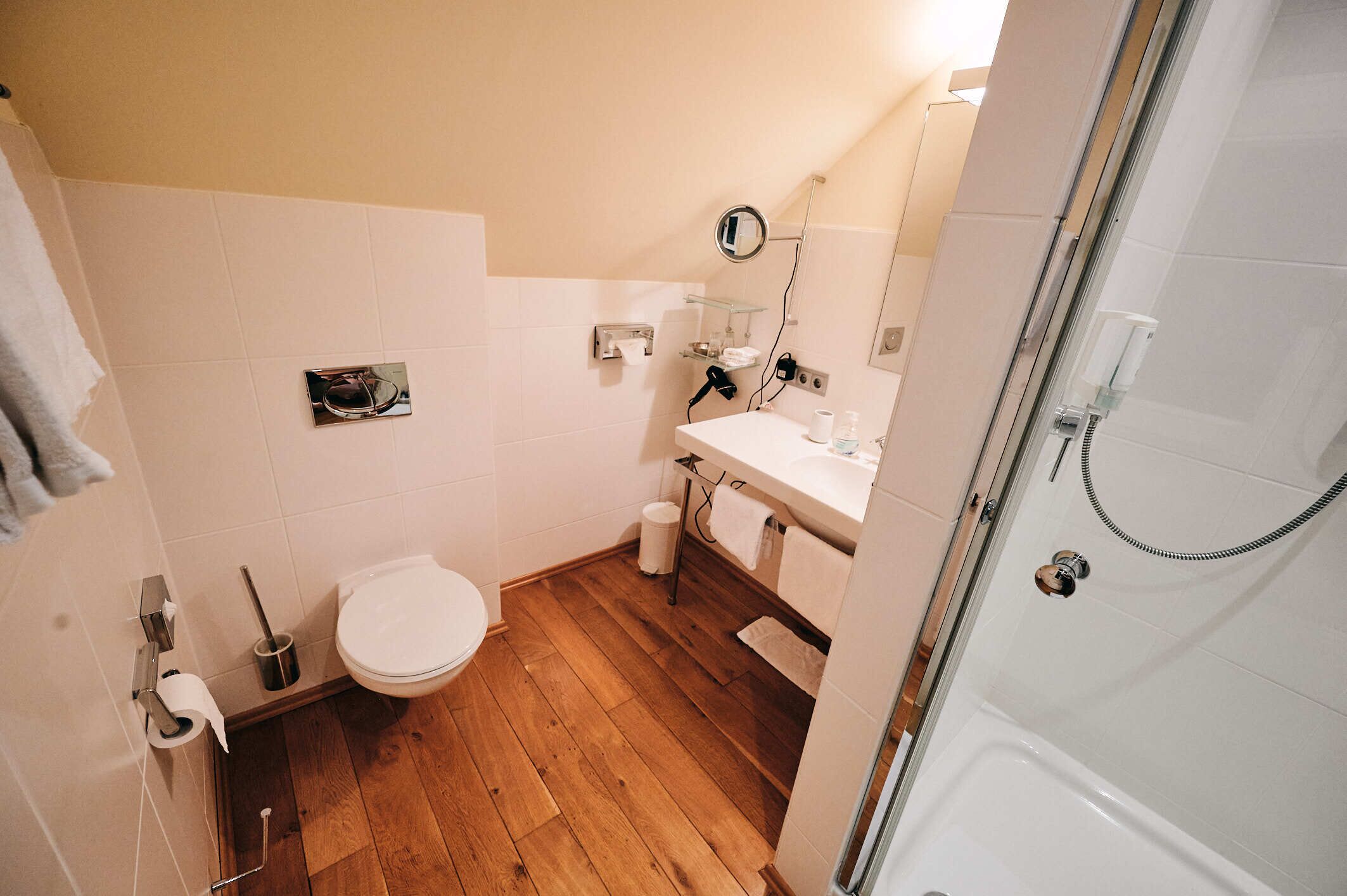 Ein Badezimmer mit Holzboden, weißen Fliesen, einer Toilette, einem Waschbecken mit Spiegel und einer Dusche.