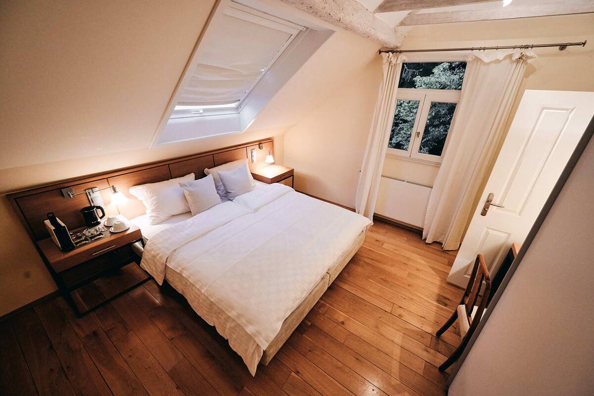 A bedroom with a skylight, wooden floor, a bed with white linens, a bedside table with a lamp, and a window with curtains.