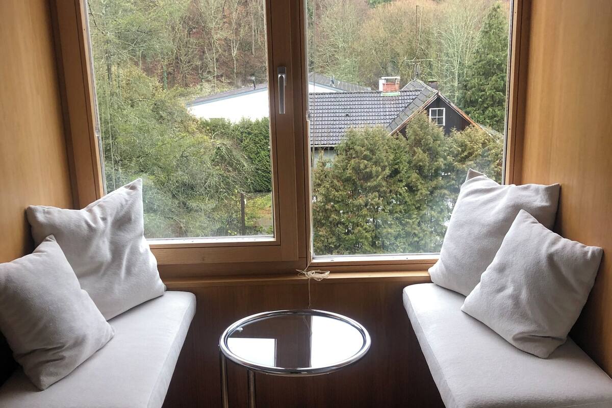 A corner seating area with two cushioned benches and a small round table, overlooking a forested area with a house in the background.