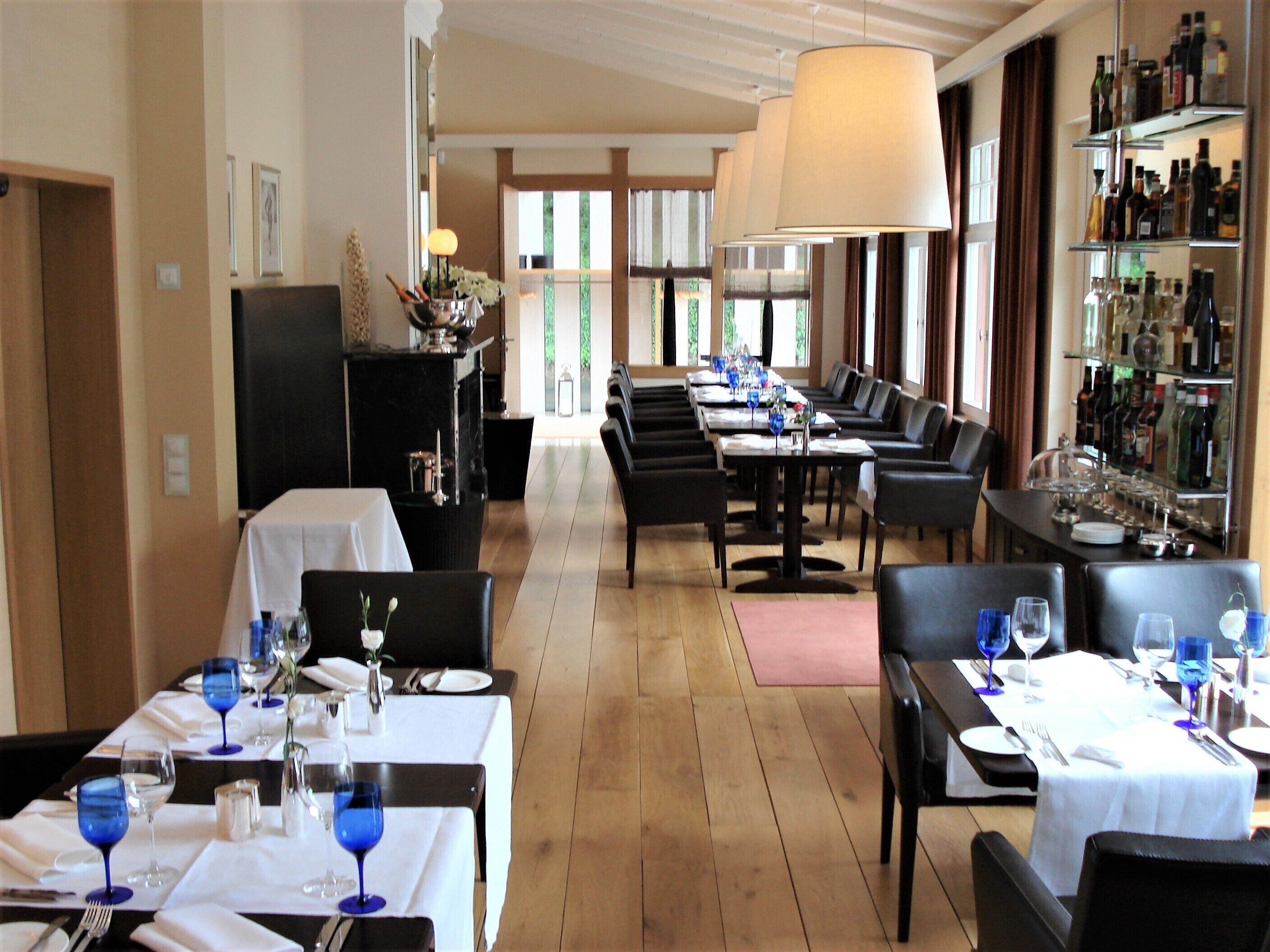 Ein Restaurant mit mehreren gedeckten Tischen für Gäste, einer Bar mit einer Auswahl an Flaschen und Blick ins Freie durch große Fenster.