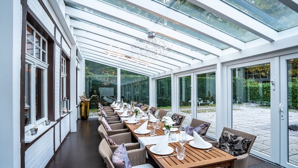 Un salón de invernadero con una larga mesa de comedor dispuesta para una comida, rodeada de sillas de mimbre.