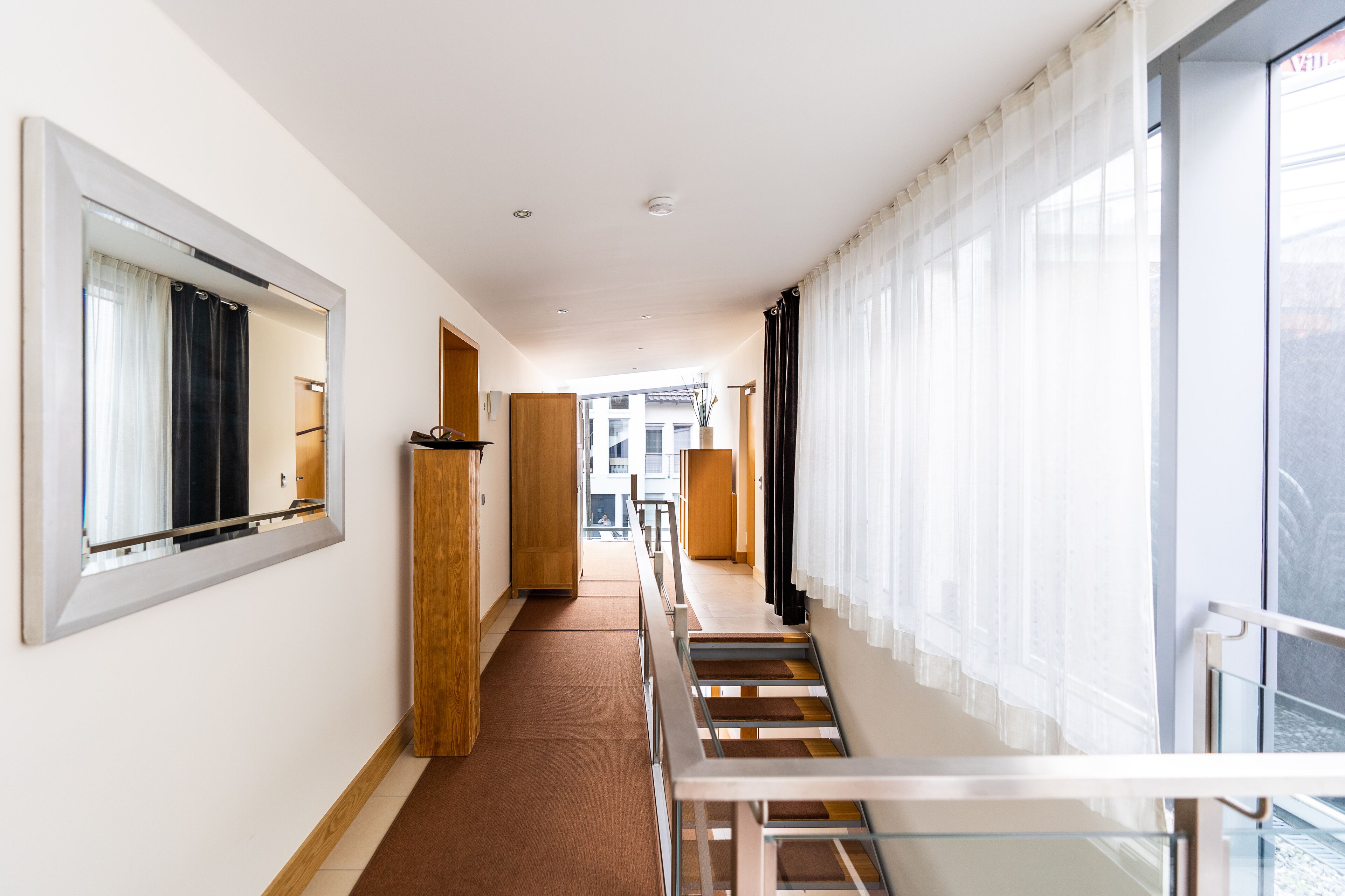 Ein Hotelgang mit Holzboden, Glastrennwand und großen Fenstern mit transparenten Vorhängen.