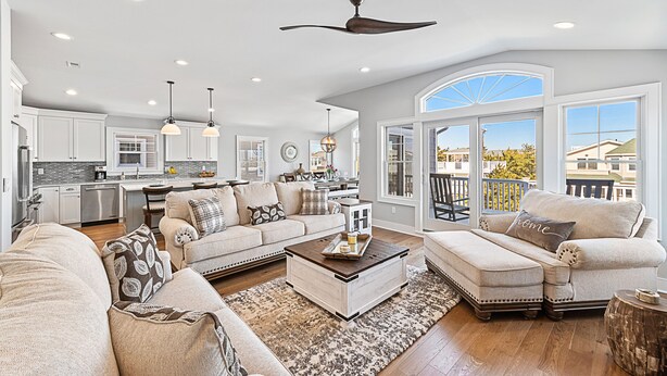 A spacious living room with a large window, a ceiling fan, and a kitchen in the background. - NEW LUXURY HOME, 2 houses from BEACH,5 Bedroom,Sleeps 12,Heated POOL, Game Room (Long Beach Township)