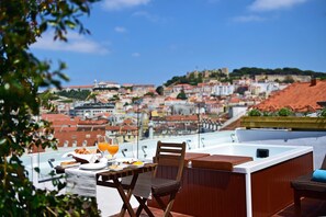 A rooftop terrace with a table set for breakfast, a hot tub, and a scenic view of a city with red-roofed buildings. - Casa Balthazar (Lisbon)