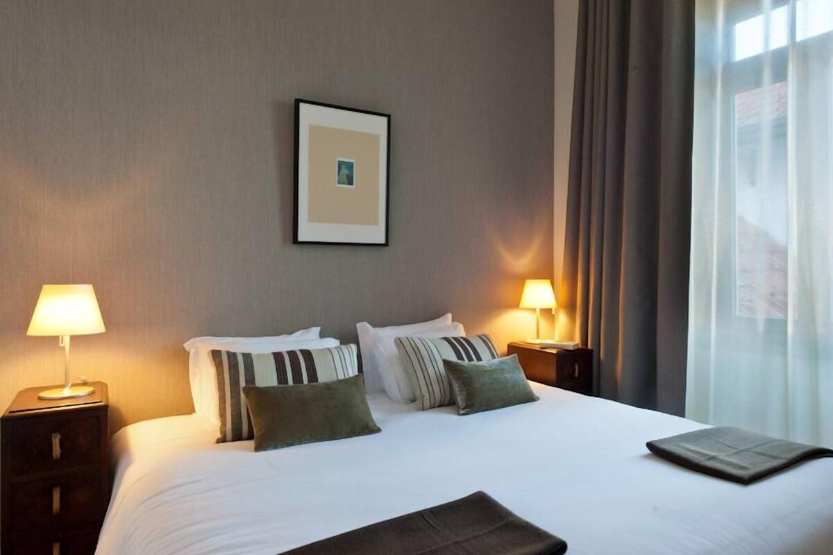 Una cama bien hecha con sábanas blancas y varias almohadas, dos lámparas de noche, un cuadro en la pared y una ventana con cortinas.