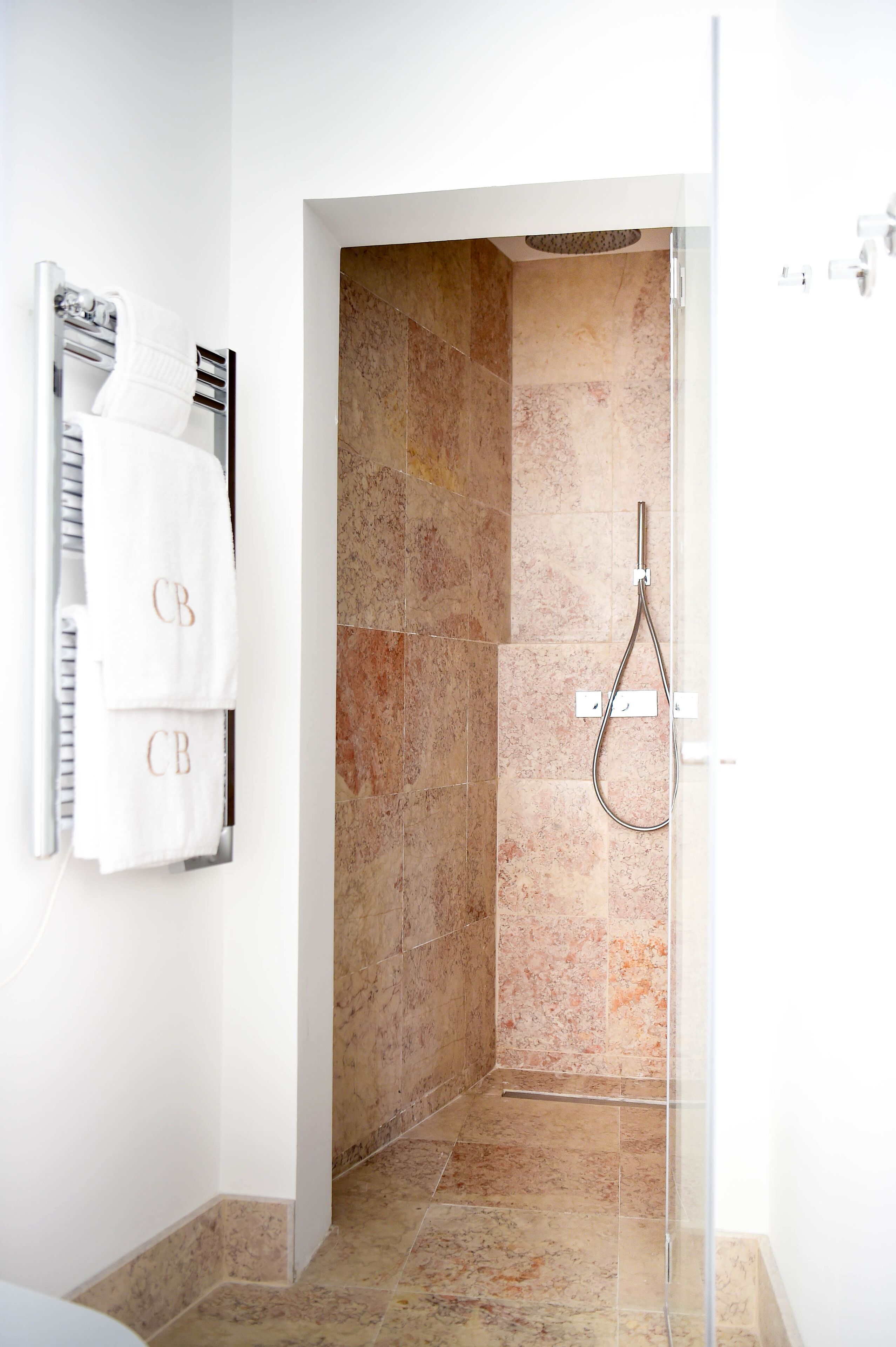 A modern bathroom with a glass-enclosed shower, beige marble tiles, and two white towels with "CB" monogrammed on them.