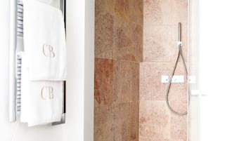 A modern bathroom with a glass-enclosed shower, beige marble tiles, and two white towels with "CB" monogrammed on them.
