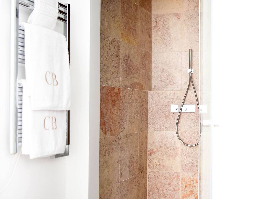 A modern bathroom with a glass-enclosed shower, beige marble tiles, and two white towels with "CB" monogrammed on them.