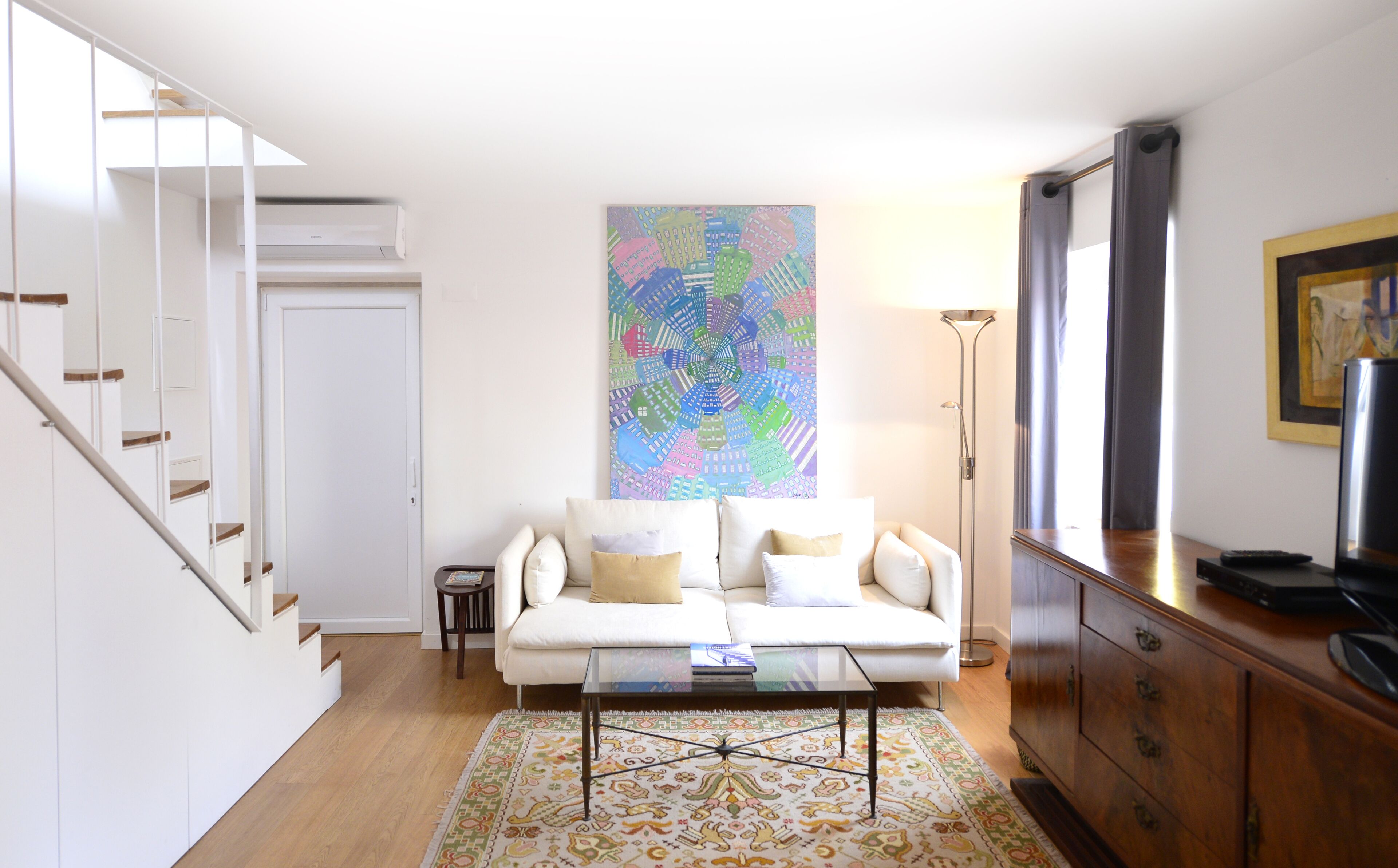 Una sala de estar con un sofá blanco, una mesa de centro de vidrio, un cuadro abstracto colorido, una lámpara de pie y un mueble de madera.