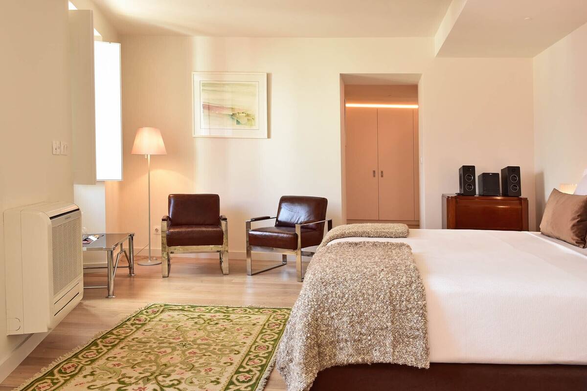 Habitación de hotel con una cama, dos sillas, una lámpara de pie, un radiador, un tapete verde estampado, una mesita de noche de madera y un altavoz montado en la pared.