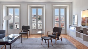 Un salon moderne avec une table basse en verre, deux fauteuils, un canapé et un téléviseur posé sur une étagère. De grandes fenêtres offrent une vue sur la ville.