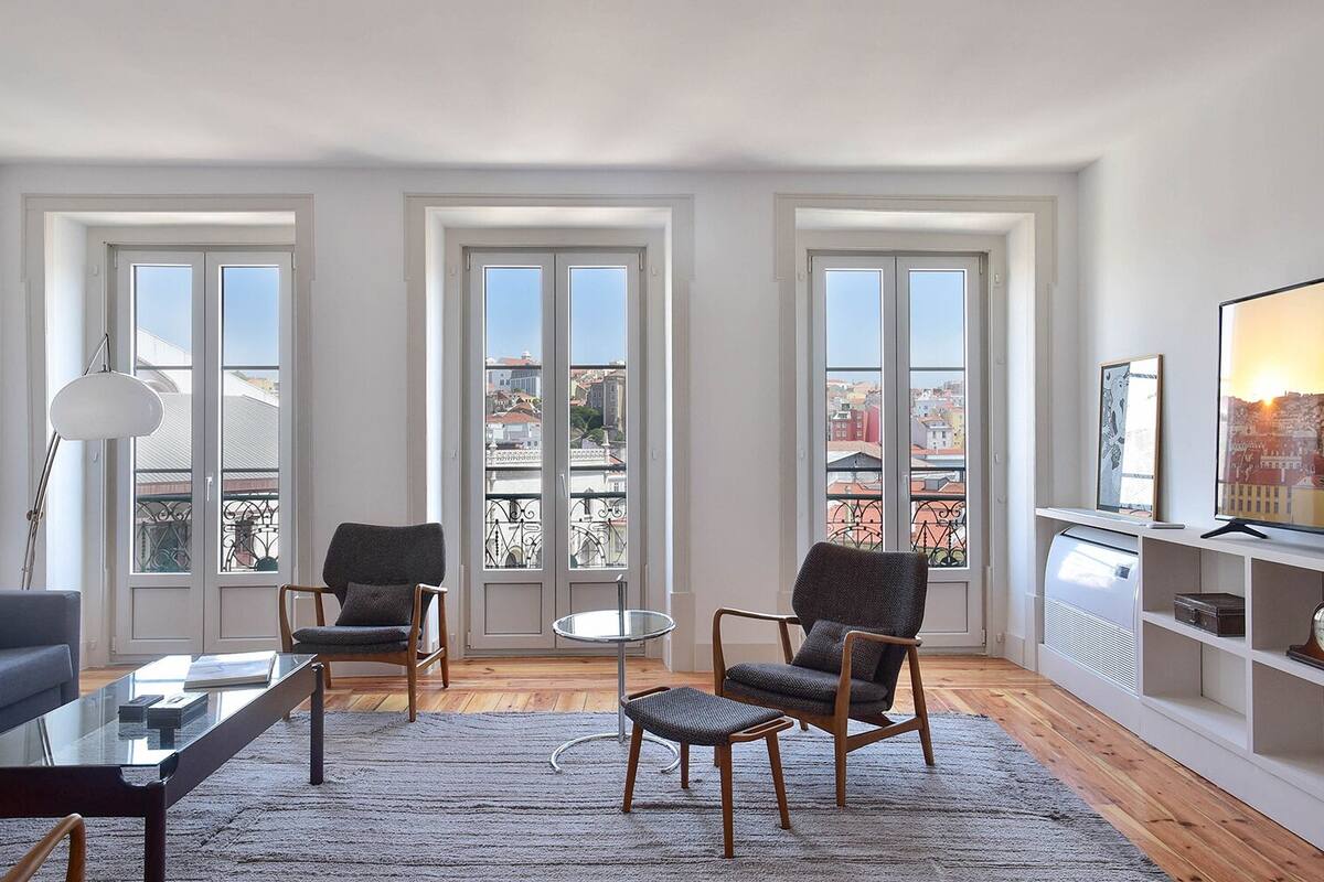 Una sala de estar moderna con una mesa de centro de vidrio, dos sillones, un sofá y un televisor en una estantería. Amplias ventanas ofrecen vistas a la ciudad.