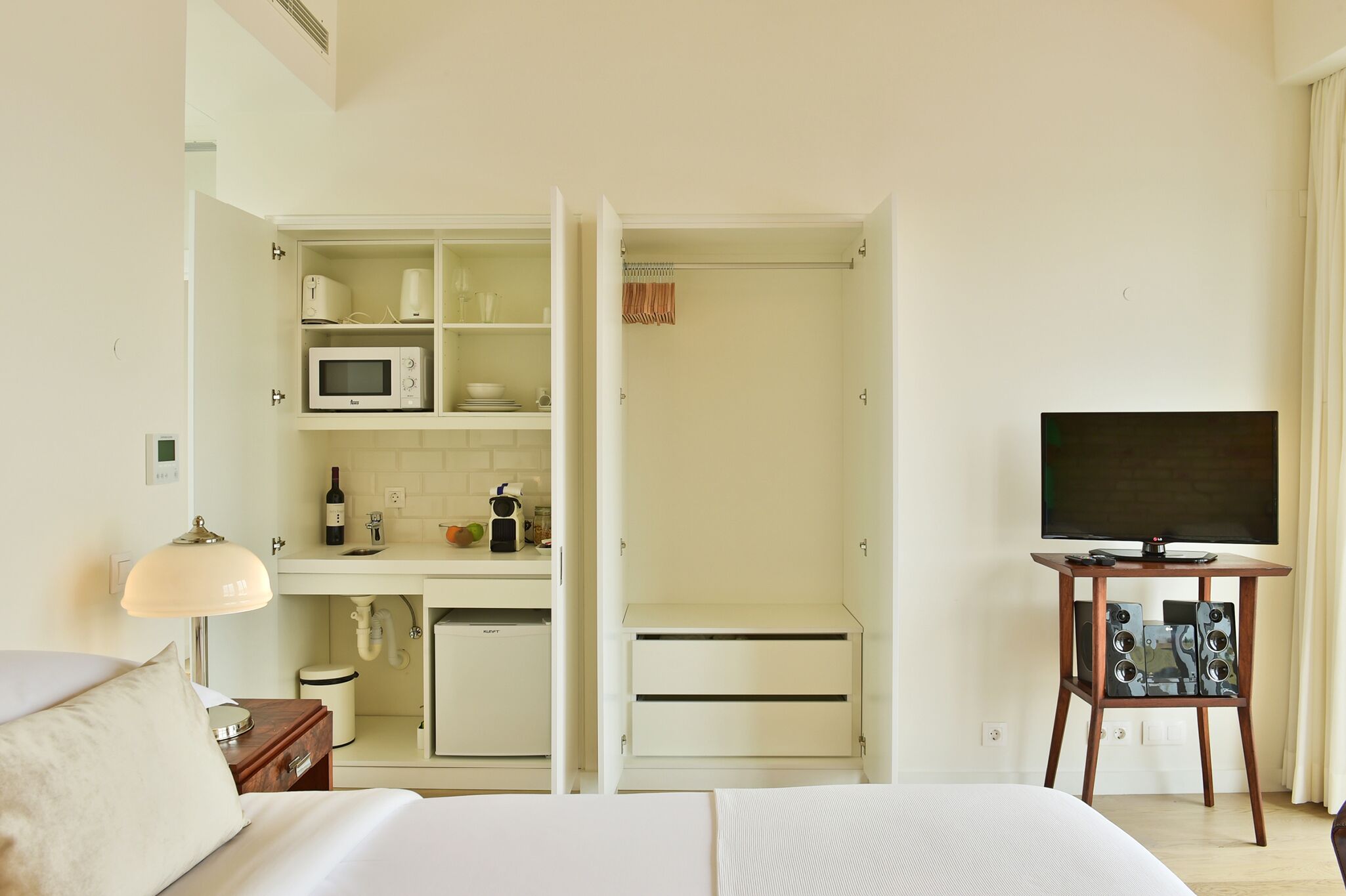 A hotel room with a bed, a TV on a stand, a kitchenette with a microwave, and a small cabinet.
