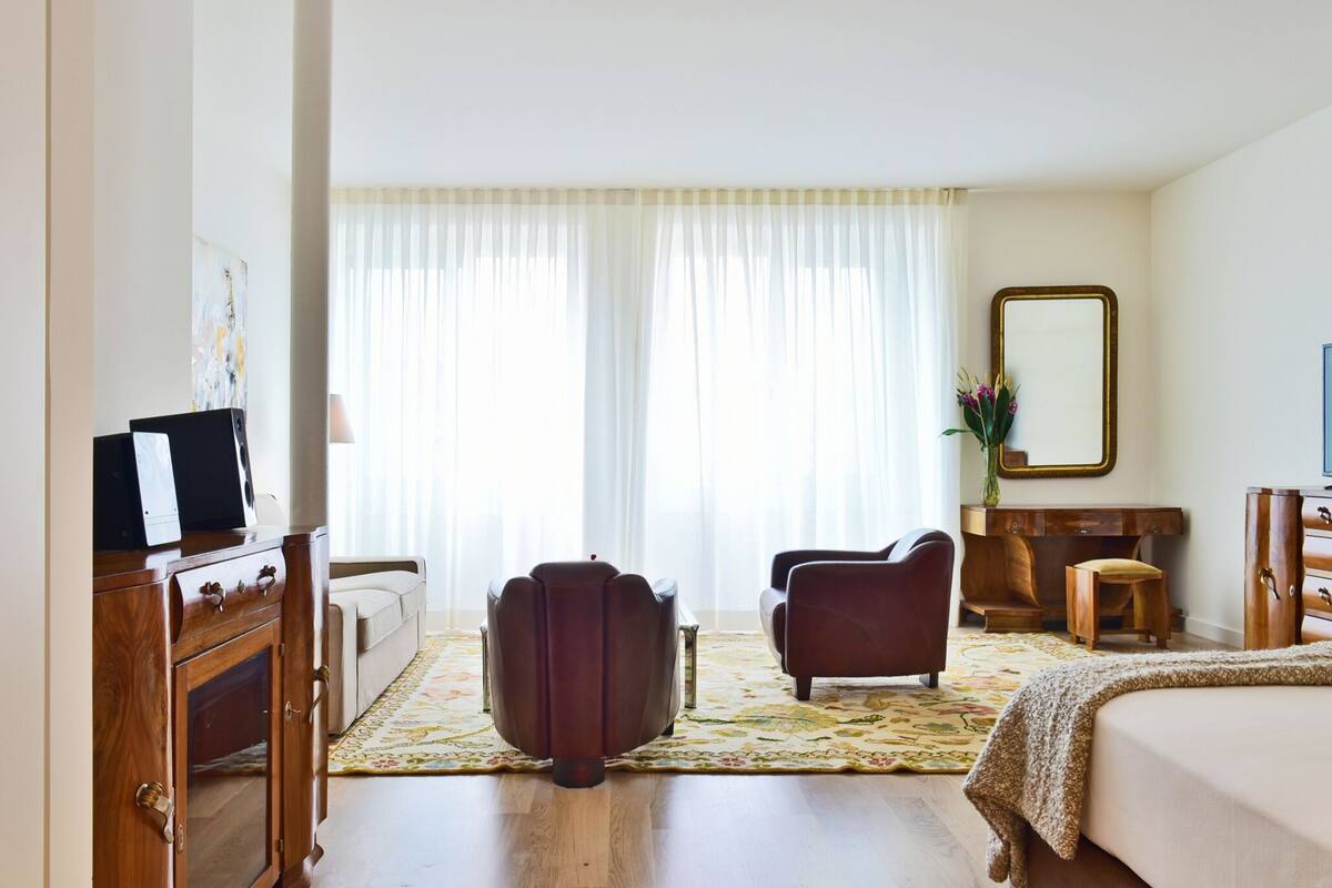 Una habitación de hotel con un aparador de madera, un sillón de cuero, un taburete pequeño, una cama, una mesita de noche de madera y un espejo en la pared.