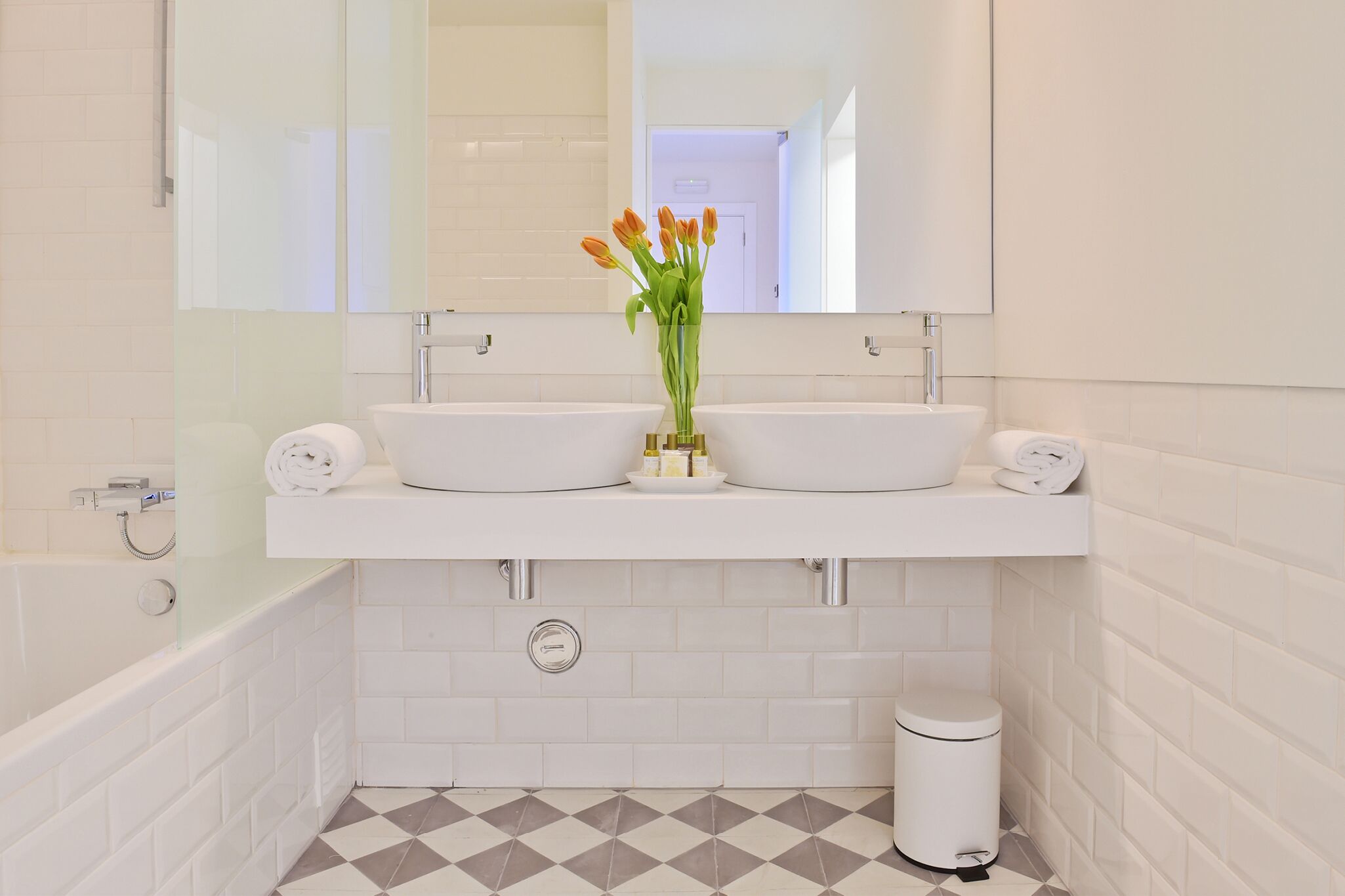 Un baño con un lavamanos blanco, dos dispensadores de toallas, un jarrón de cristal con flores, un espejo y un suelo de baldosas.