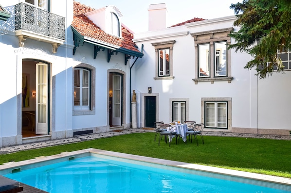 A poolside area with a dining table and chairs, a small statue, and a well-maintained lawn. - Casa Balthazar (Lisbon)