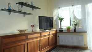 A living room with a TV on a wooden cabinet, decorative shelves, a window with curtains, and a radiator. - Vacation apartment Haus am Jakobsweg (Fischbach bei Dahn)