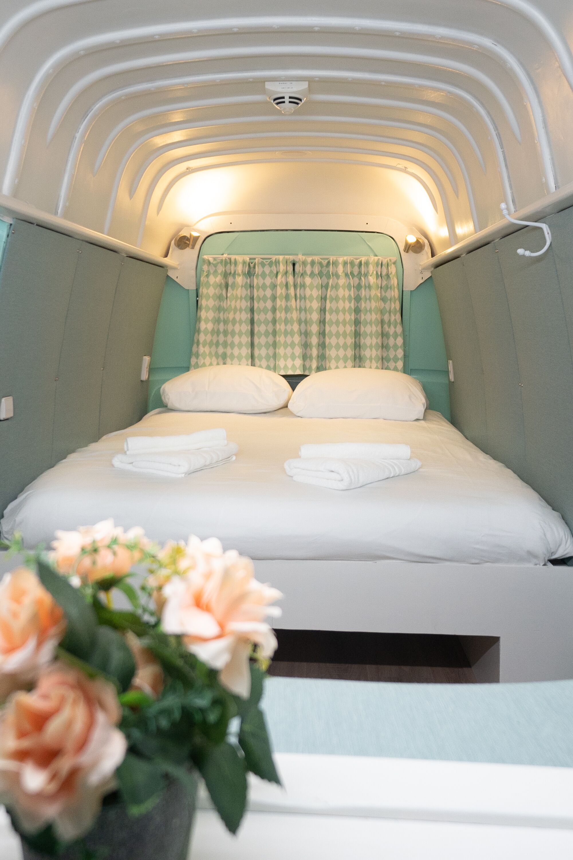 Un dormitorio compacto y bien iluminado, con una cama bien hecha, una cortina a cuadros y un jarrón con flores.