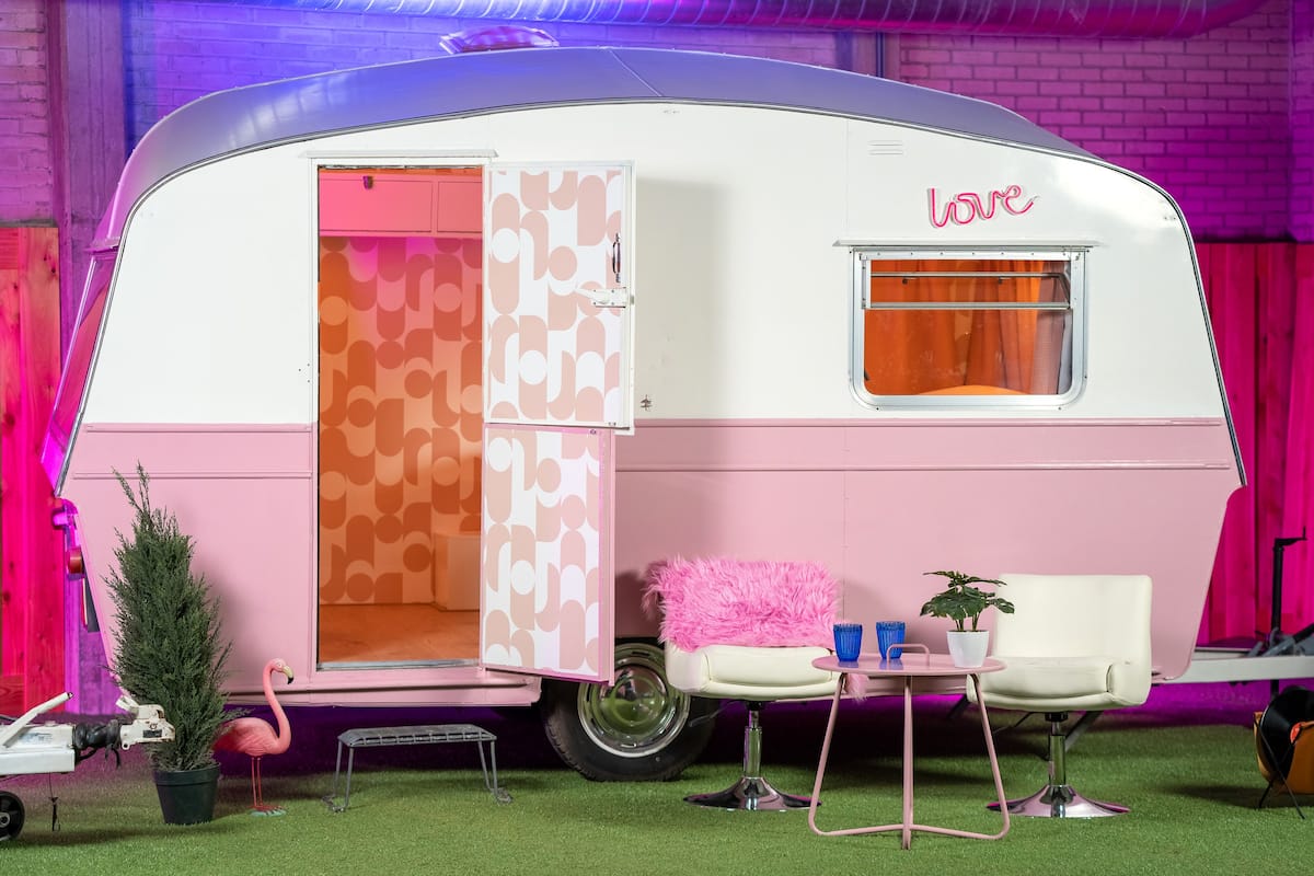 A pink and white trailer with a 'love' sign, a small table with chairs, a potted plant, and a flamingo statue.