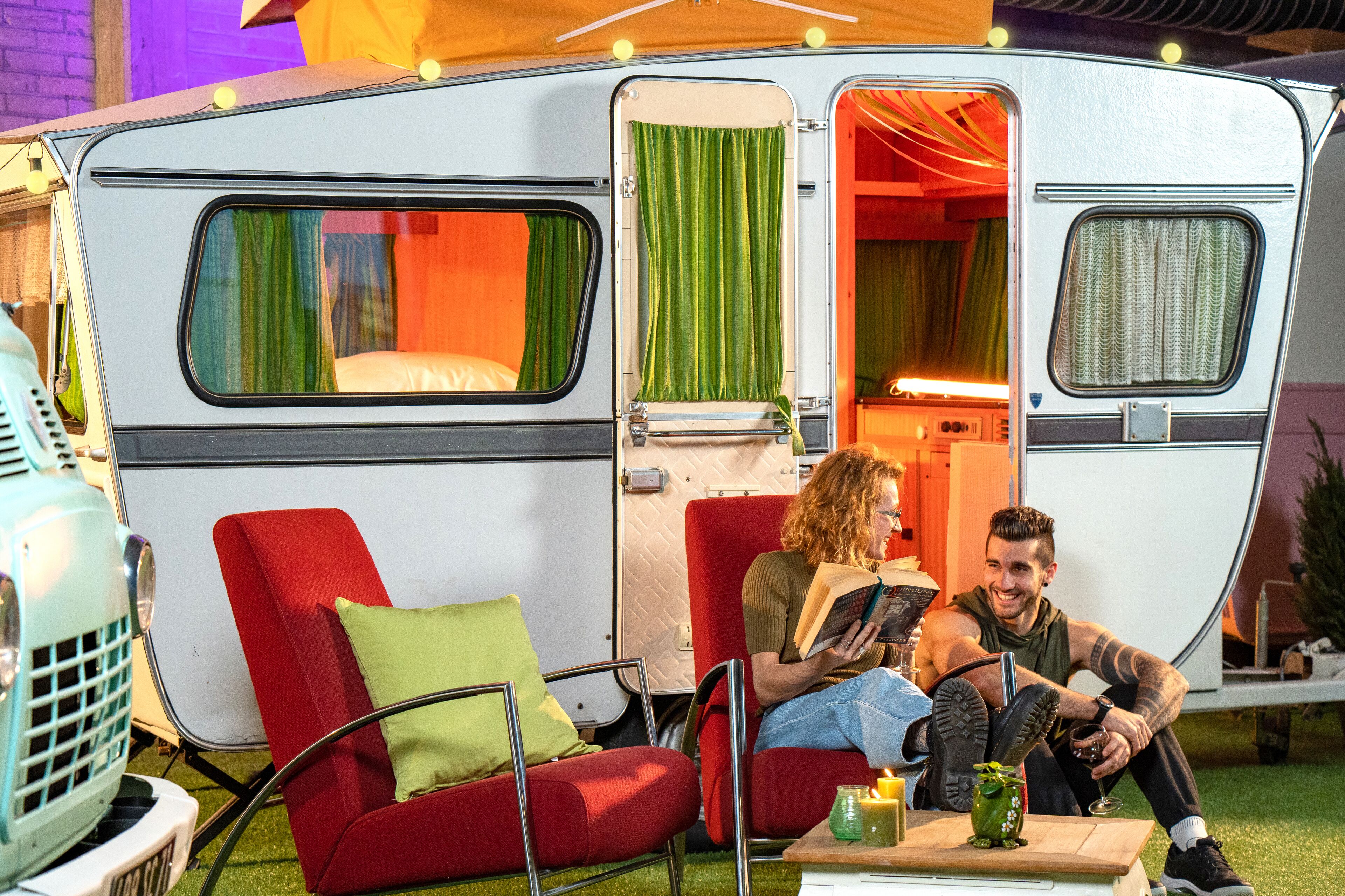 A camper van interior with a red sofa, a small table with a drink, and a person reading a book.