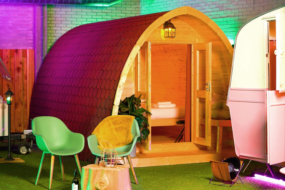 A cozy, curved wooden structure with a red roof, a small table with a bottle, two chairs, and a potted plant.