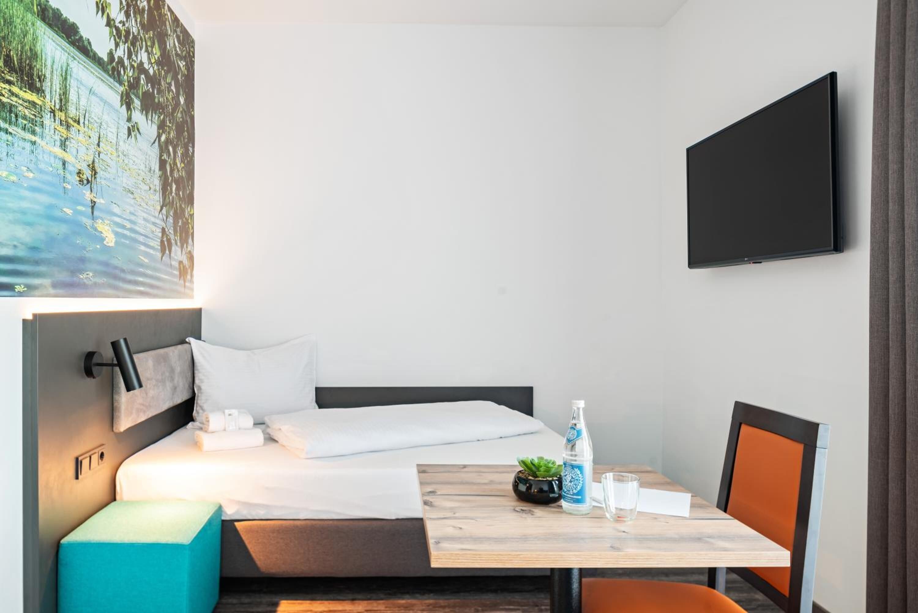 Habitación de hotel con cama, una mesa con una botella y un vaso, un televisor en la pared y un cuadro de un lago.