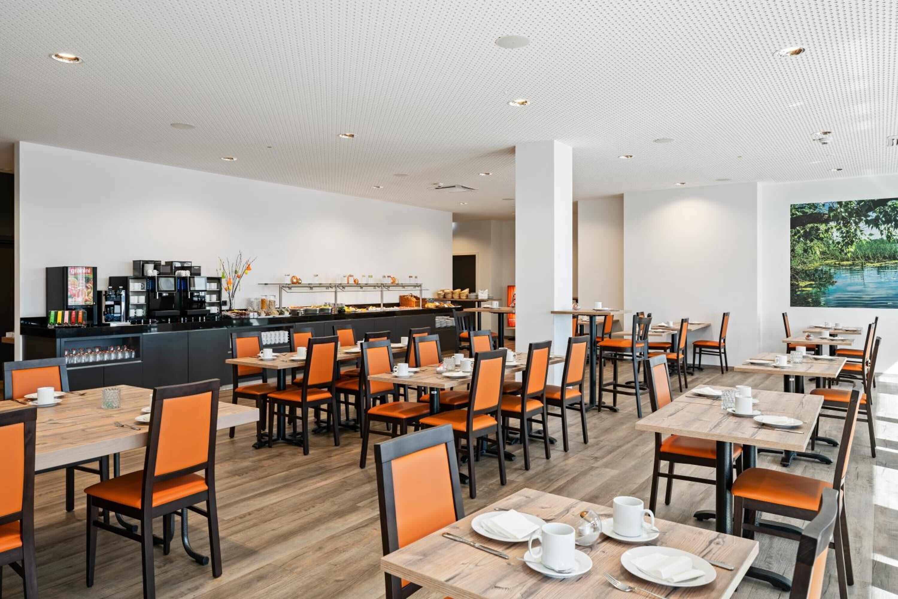 A modern dining area with multiple tables and chairs, a kitchen area with appliances, and a large window with a view of greenery.