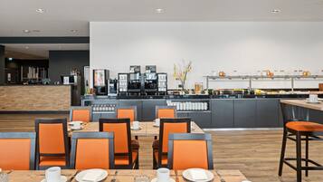 A modern dining area with orange chairs, wooden tables, and a kitchen in the background.